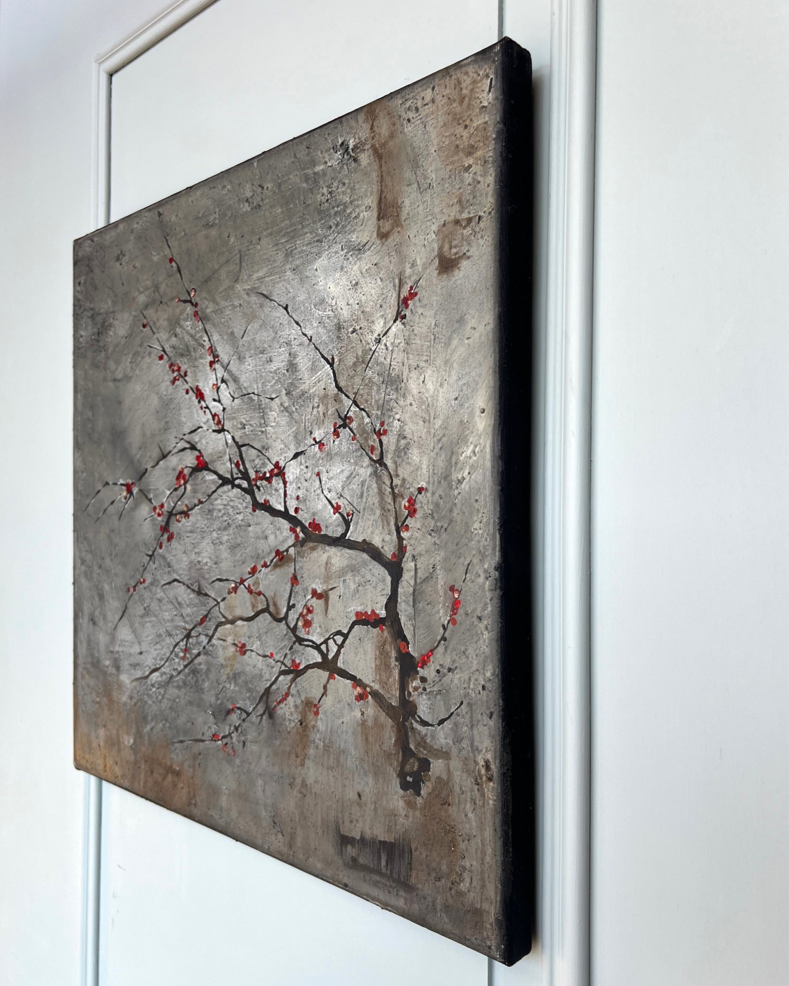 Angled view of a square contemporary painting with visible canvas depth and a restrained branch motif on a weathered grey surface.