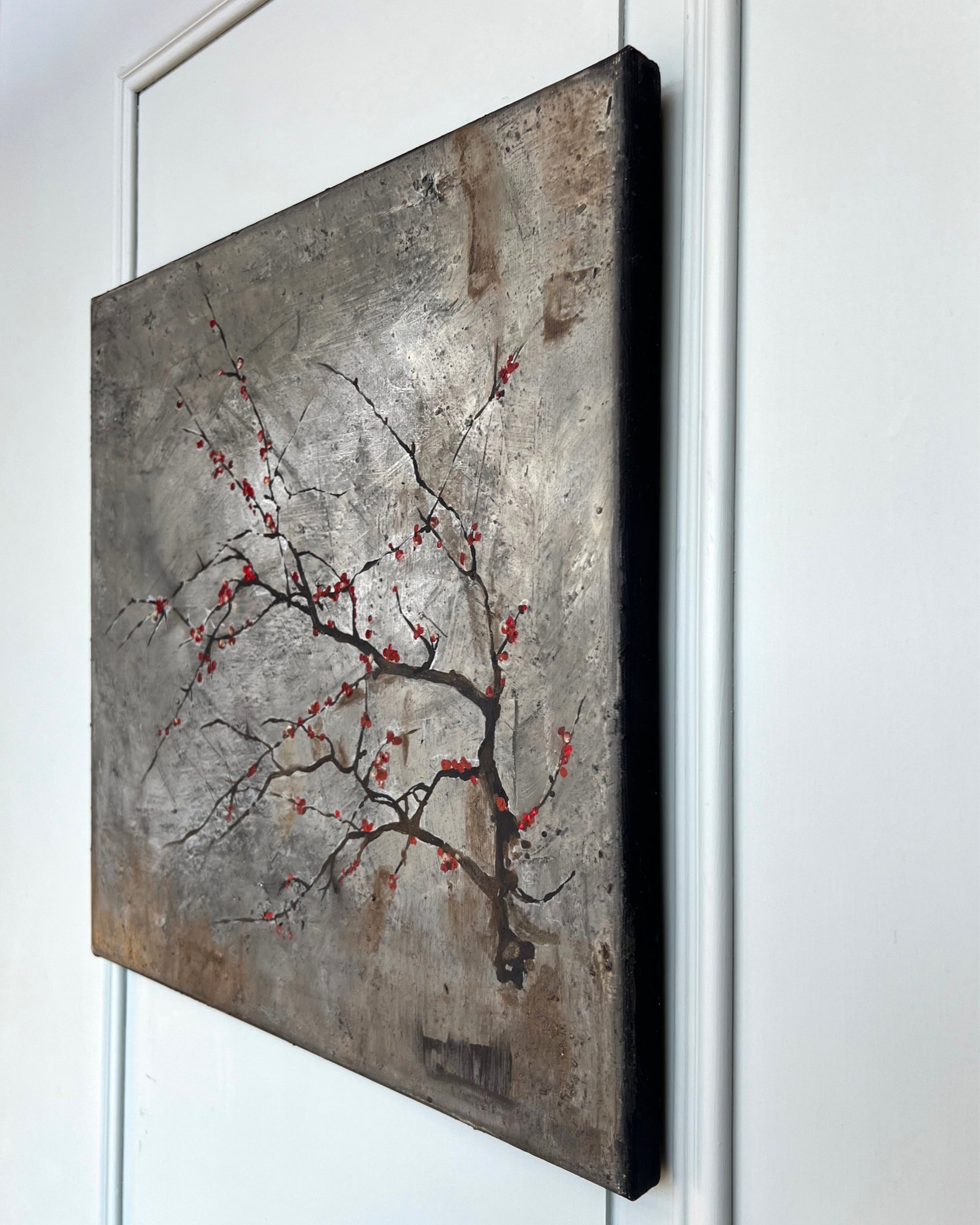 Angled view of a square contemporary painting with visible canvas depth and a restrained branch motif on a weathered grey surface.