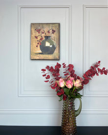 Small still life artwork displayed in a calm interior with floral arrangement and neutral wall panels.